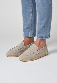 Beige suède instapschoenen met een gestikt ontwerp, voorzien van een geweven jute zool en een platte hak. Geschikt voor casual gebruik.