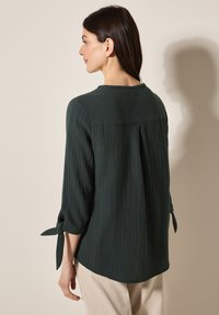Top texturizado verde oscuro con mangas tres cuartos, con detalles de lazo en los puños y un dobladillo ligeramente curvado.