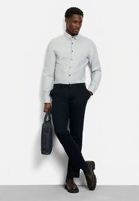 Hellgraues Hemd mit Knöpfen in Dunkel, schwarze Hose und braune Schuhe. Trägt eine dunkle Tasche, steht in neutraler Haltung.