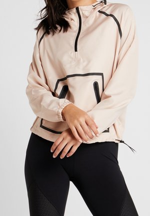 Femme portant une veste beige à demi-fermeture éclair avec une fermeture noire et des contours de poches, associée à des leggings noirs avec des panneaux texturés.