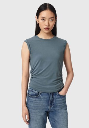 Femme aux cheveux lisses et foncés porte un haut bleu sans manches et un jean bleu, debout avec une main dans la poche devant un fond clair.