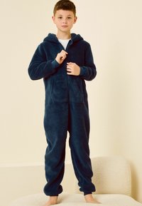 Marineblauwe fleece onesie met een ritssluiting aan de voorkant, een capuchon en lange mouwen. Bevat zijzakken en ribbelmanchetten. Zachte, gestructureerde oppervlakte.