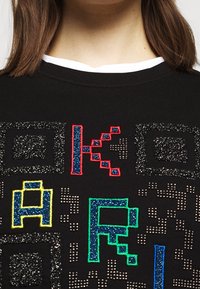 Černá mikina s barevnými, třpytivými vyšívanými písmeny a vzory v červené, modré, zelené, žluté a stříbrné textuře.