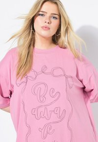 Rosa Übergröße T-Shirt mit kurzen Ärmeln, das eine schwarze Linienzeichnung des Satzes "Sei achtsam" und dekorative Schleifen zeigt. Weicher Baumwollstoff.