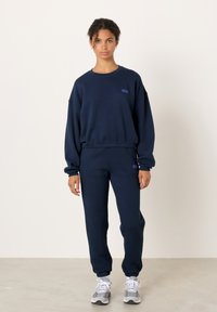 Tamsiai mėlynas džemperis ir atitinkamos kelnės iš minkšto medvilnės, su gofruotais rankogaliais ir mažu logotipu. Paprastas dizainas su laisvu kirpimu.