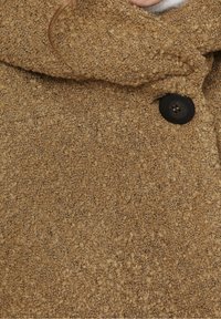 Abrigo texturizado marrón con un gran cuello, cierre de botones negros y un patrón de tela tejida. Acabado suave con una apariencia cálida.
