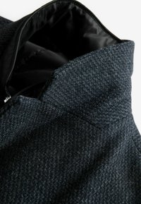 Wollmischungs-Mantel mit einem strukturierten, dunkelgrauen Muster. Verfügt über einen Reverskragen und ein schwarzes Futter. Nahaufnahme zeigt die Stoffdetails.