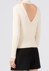 Cremfarbener Strickpullover mit hohem Kragen, langen Ärmeln und einer tiefen, V-förmigen Rückenöffnung. Geripptes Bündchen und Manschetten. Getragen mit schwarzen Shorts.