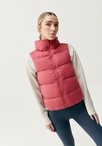 Röd pufferväst med hög krage och framdragkedja, med horisontella quiltade paneler. Bärs över en ljusfärgad långärmad tröja och blå leggings.