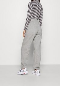 Mujer en pantalones de chándal grises, zapatillas blancas y plateadas, y una camiseta de rayas blancas y negras de manga larga, de espaldas sobre un fondo sencillo.