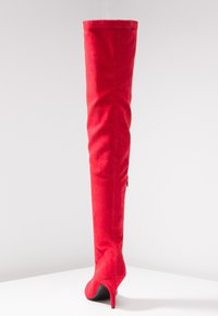 Botte rouge cuissarde en daim doux avec un design élégant, un bout pointu et un talon aiguille. Détail de fermeture éclair latérale sur la tige inférieure.