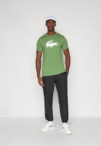 Zaļš kokvilnas T-krekls ar baltu krokodila logo, apvienots ar melnu biksēm un baltajiem sporta apaviem. Modelis valkā baltu cepuri.