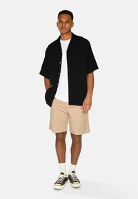 Cămașă neagră cu mâneci scurte peste un tricou alb, asociată cu pantaloni scurți bej. Tenisi negri cu șireți albi și șosete scurte completează look-ul.