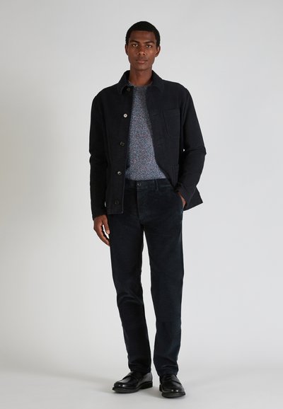 Navy Cord-Jacke mit Knopfverschluss, kombiniert mit einem mehrfarbigen strukturierten Pullover und dunklen Hosen. Schwarze Lederschuhe runden das Outfit ab.