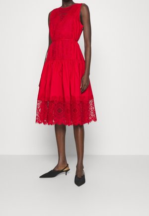 Kobieta w czerwonej sukience bez rękawów z koronkowymi detalami i rozszerzanym dołem, w połączeniu z czarnymi czółenkami na niskim obcasie z odkrytą piętą i spiczastym noskiem.