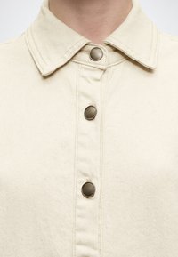 Γιακάς πουκάμισου από μπεζ τζιν με τρία ορατά μεταλλικά κουμπιά τύπου snap κλεισμένα στην μπροστινή λάστιχη.