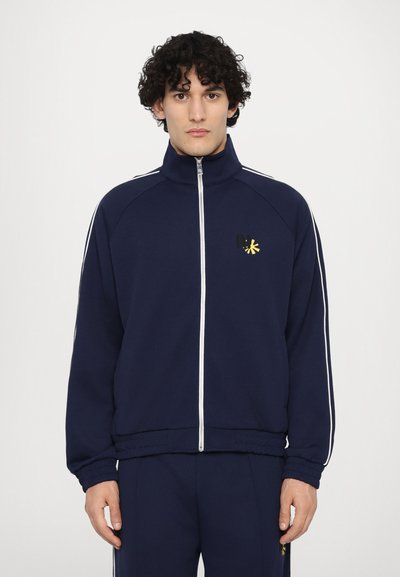 Veste de survêtement zippée bleu marine avec des rayures blanches, présentant un petit logo brodé. Tissu doux, les poignets et l'ourlet ribbed apportent de la structure.