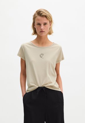 Femme aux cheveux blonds courts portant un t-shirt beige avec un petit motif cœur et un pantalon noir à cordon, debout les mains dans les poches.