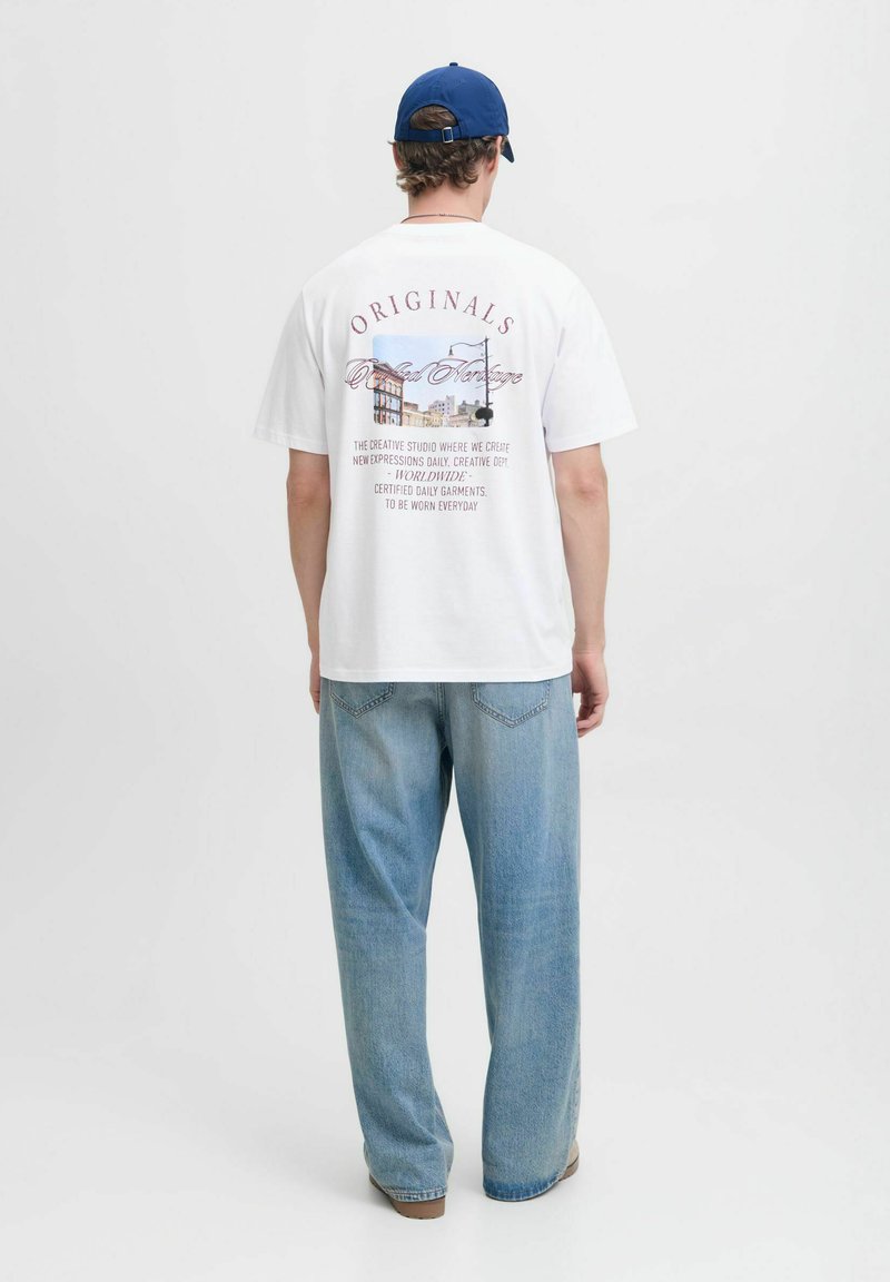 Maglietta bianca a maniche corte con una stampa grafica sul retro, con testo in diversi caratteri e colori. Abbinata a jeans a gamba larga di colore blu chiaro.