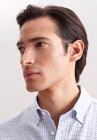 Camicia button-up a quadri blu chiaro e bianca con colletto, realizzata in tessuto morbido. I corti capelli scuri del modello e i tratti delicati del viso sono visibili.