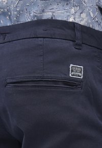 Temno modre bombažne hlače imajo zadnji žep z majhno, belo obrobljeno etiketo z napisom "GUESS Est. 1981" v črni pisavi.