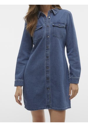 Kvinde iført en blå denim kjole med knapper foran, lange ærmer og to brystlommer, stående mod en ensfarvet baggrund.