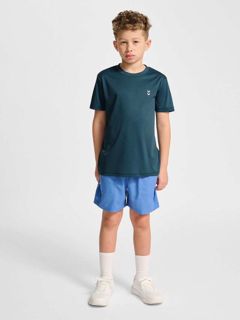 Chłopiec z krótkimi kręconymi włosami, ubrany w ciemnozielony t-shirt, jasnoniebieskie krótkie spodenki, białe skarpetki i białe trampki, stojący na jednolitym tle.