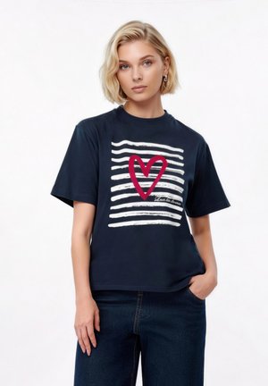 Blondynka w granatowej koszulce z białymi poziomymi paskami i czerwonym sercem na przodzie, w ciemnoniebieskich dżinsach, stojąca z jedną ręką w kieszeni.