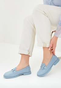 Lichtblauwe loafers van Perforatiemateriaal met witte stiksels en een strikdetail aan de voorkant, gecombineerd met opgerolde witte broeken.