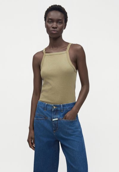 Femme aux cheveux courts et bouclés portant un haut beige sans manches à encolure carrée et un jean bleu à jambes larges, main dans la poche, fond neutre.