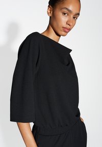 Top nero a maniche lunghe con scollatura tonda e vestibilità comoda. Realizzato in tessuto texturizzato, presenta un dettaglio con cordoncino in vita.