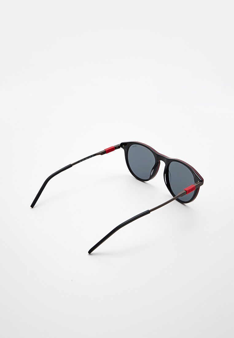 Occhiali da sole con montatura rotonda in plastica nera, lenti scure e dettagli rossi sulle aste. Design elegante e leggero.