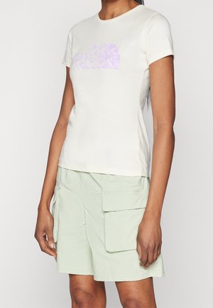 Kobieta ubrana w białą koszulkę z krótkim rękawem z delikatnym fioletowym logo oraz jasne zielone szorty typu cargo z dużymi przednimi kieszeniami.