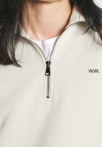 Světlé béžové mikina na zip s texturovanou tkaninou, stříbrným detailem zipu a malým vyšívaným logem "W.W." na hrudi.