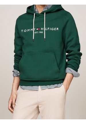 Mężczyzna w ciemnozielonej bluzie z kapturem Tommy Hilfiger na koszuli w kratę i beżowych spodniach, z jedną ręką w kieszeni i widoczną przednią kieszenią kangurzą.