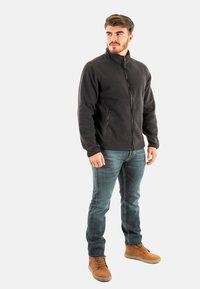 Chaqueta de forro polar negra con cuello alto y cremallera frontal, con dos bolsillos laterales. Llevada con jeans de mezclilla azul y botas marrones.