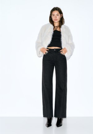 Čierne džínsy s širokými nohavicami so štýlovým detailom s cvokmi na boku, skombinované s bielou bundou z umelého kožúšku a čiernym krátkym topom. Modelka má na sebe čierne členkové čižmy na opätku.