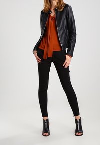 Schwarze Lederjacke mit taillierter Passform, orangefarbenes Oberteil mit gebundenem Vorderteil, schwarze schmal geschnittene Hose und offene schwarze Stiefeletten.