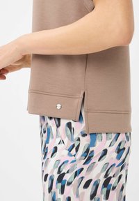 Braunes, leichtes Oberteil mit seitlichen Schlitzen; silberne Hardware-Details am Saum. Tragen mit gemusterten Hosen, die Pastell-Pinselstrich-Designs aufweisen.