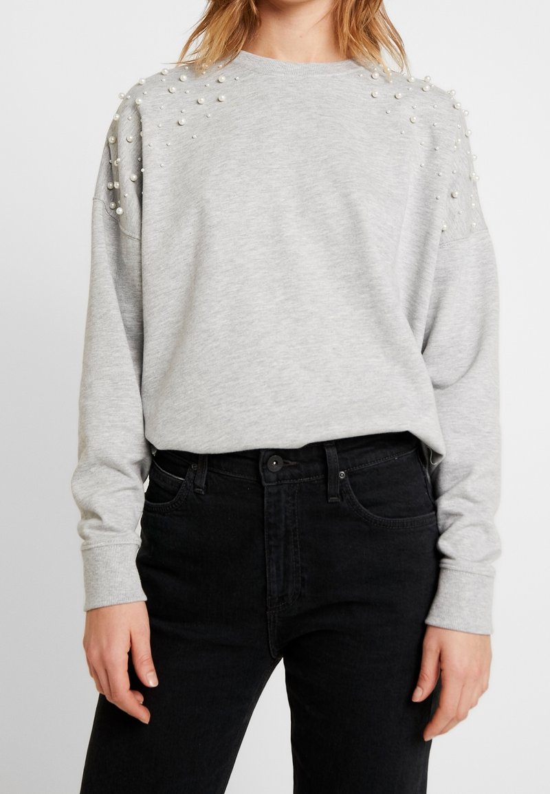 Sweat-shirt gris avec des perles ornant les épaules, des poignets et un ourlet côtelés. Porté avec un jean noir, illustrant une tenue décontractée.