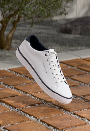 Hvid Tommy Hilfiger sneaker med sort kant og snørebånd, svævende over trægulve og små grå sten udendørs.