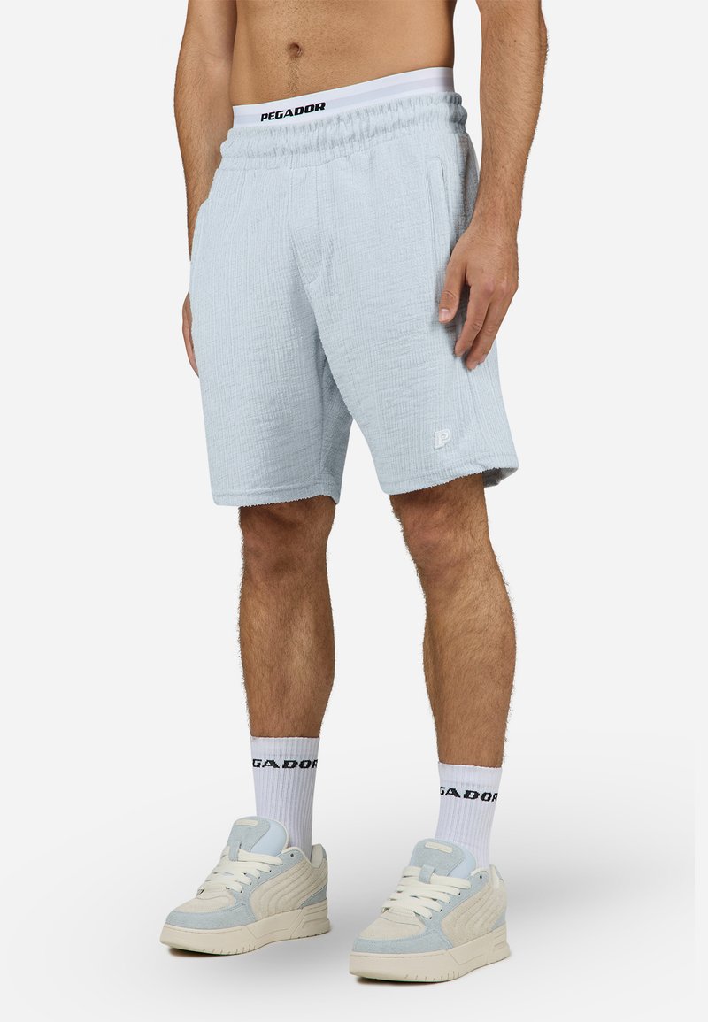 Pantaloni scurți texturați de culoare albastru deschis, cu talie elastică și buzunare laterale, având un logo mic pe tiv. Asortați cu șosete albe cu logo și adidași deschiși la culoare.