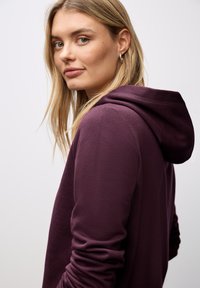 Sudadera con capucha morada de textura suave, con mangas largas y un corte relajado. Presenta una capucha y un notable detalle de costura.