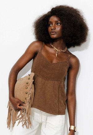 Femme aux grands cheveux bouclés porte un haut marron sans manches, un pantalon blanc, un collier en or, un bracelet et tient une pochette marron à franges avec des clous.