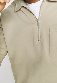 Personne portant un pull à fermeture éclair beige sur une chemise blanche, avec une poche sur le côté gauche de la poitrine et la fermeture éclair partiellement ouverte.