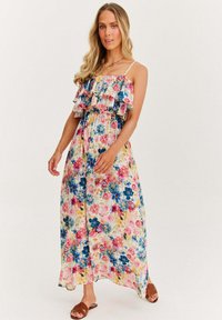 Kwiatowa sukienka maxi z cienkimi ramiączkami, z falbaną przy dekolcie, ozdobiona różowymi i niebieskimi kwiatami na jasnym tle. Brązowe sandały.