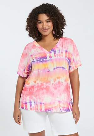 Kobieta z kręconymi włosami w różowej, pomarańczowej i fioletowej koszulce batikowej z dekoltem w serek oraz białych szortach, stojąca na jasnym, jednolitym tle.