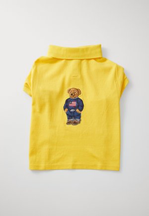 Polo amarelo de algodão com um urso bordado numa jaqueta azul, calças de ganga e sapatos castanhos nas costas.