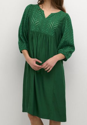 Kobieta w zielonej sukience do kolan z koronkowymi, bufkami i dekoltem, stoi z luźno splecionymi rękami.