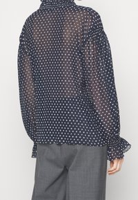 Blouse bleu marine transparente à pois blancs, manches bouffantes, texture plissée, poignets à volants et col montant. Associée à un pantalon gris.